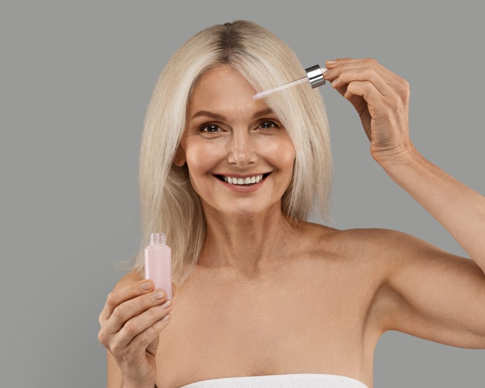 Anti-Age Cosmetics. Attractive Mature Woman Applying Face Serum With Droplet On Face, Beautiful Smiling Senior Lady Moisturising Skin While Standing Wrapped In Towel Over Grey Background
